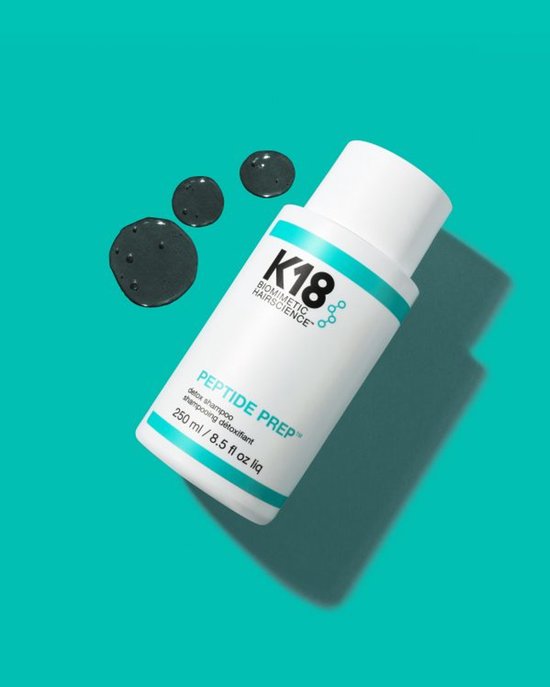 Witte fles K18 Peptide Prep Detox Shampoo met turquoise banden, gefotografeerd met drie druppels shampoo ernaast op een turquoise achtergrond.