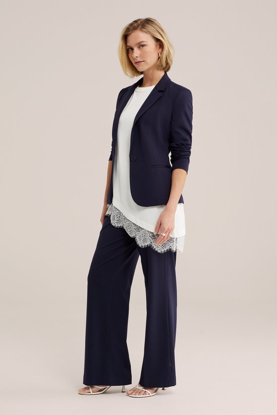Model in een donkerblauwe blazer, wit T-shirt met kanten afwerking en wijde broek.