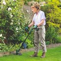 Vrouw met blauwe blouse werkt in de tuin met een Bosch grastrimmer, langs een struik met witte bloemen.