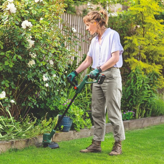 Vrouw met blauwe blouse werkt in de tuin met een Bosch grastrimmer, langs een struik met witte bloemen.