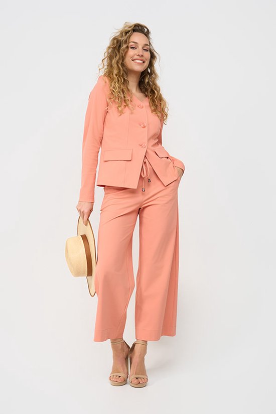 Model in een koraalkleurige broek met wijde pijpen en hoge taille, gecombineerd met een bijpassend blazerjasje. Ze houdt een strooien hoed vast.