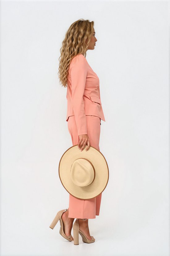Model van opzij gezien in een koraalkleurige broek met wijde pijpen en blazer. Ze houdt een strooien hoed vast.