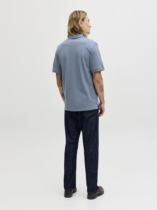 Man met blond haar en tatoeages staat met de rug naar de camera, draagt blauwgrijze polo en spijkerbroek.