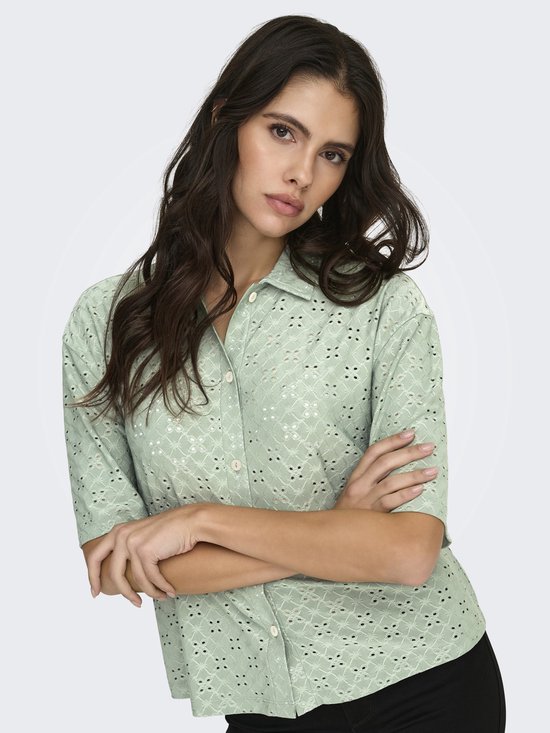Model met lichtgroene blouse met broderie anglaise, armen over elkaar.