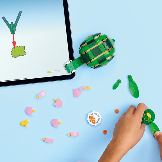 Een kind bouwt een groene legoschildpad met oranje en roze onderdelen, met een tablet en bouwinstructies ernaast.
