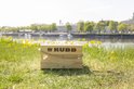 Houten Kubb kistje met touw, op gras bij een meer met gebouwen op de achtergrond.