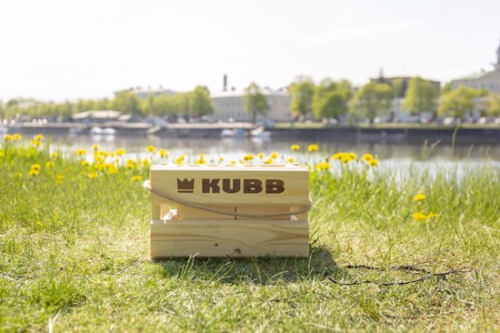 Houten Kubb kistje met touw, op gras bij een meer met gebouwen op de achtergrond.