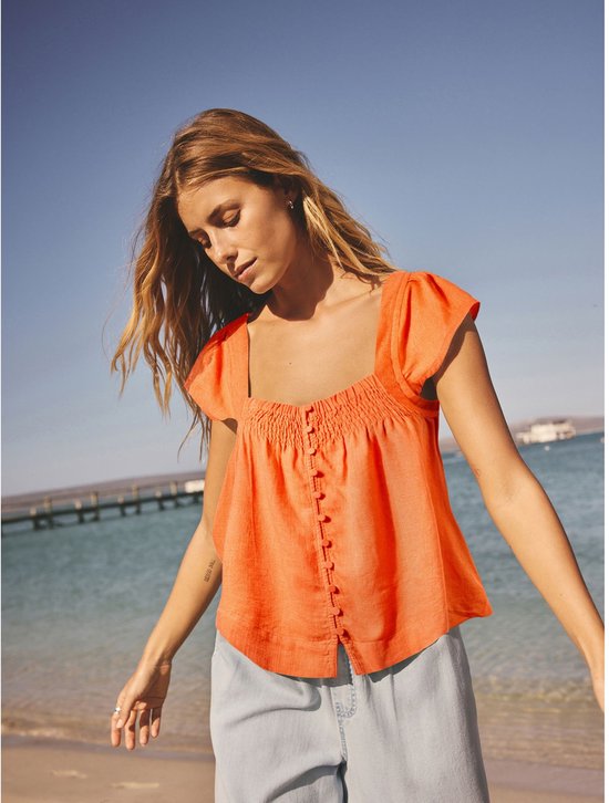 Vrouw in oranje linnen top met vierkante hals en fladdermouwen, kijkend naar beneden op een strand.