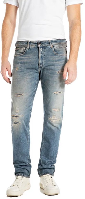 Man draagt een blauwe Replay jeans met lichte wassing en distressed details, staand.