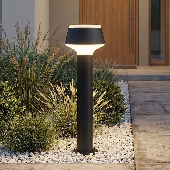 Verlichte zwarte staande lamp met conische witte kap, staand bij een entree.