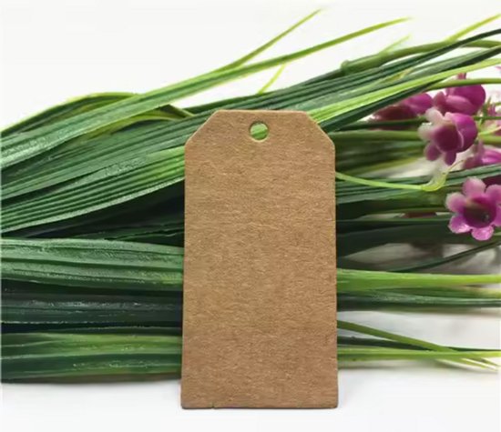 Bruin kartonnen label van 35 x 70 mm met een gat, liggend op groen gras met paarse bloemen.