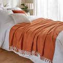 Terracotta plaid met franjes, gedrapeerd over een wit bed met kussens.