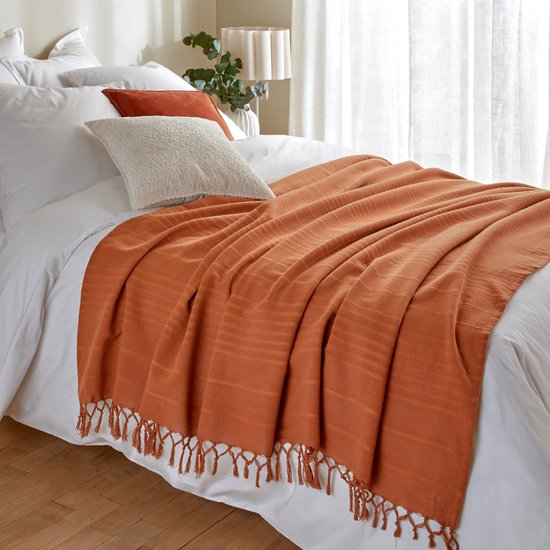 Terracotta plaid met franjes, gedrapeerd over een wit bed met kussens.