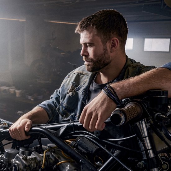 Man in spijkerjack leunt op een motor, draagt een zwarte leren armband met blauwe kralen.