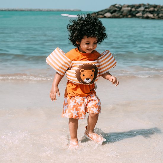Kind op strand met zwembandjes met leeuwenprint en oranje-witte strepen.