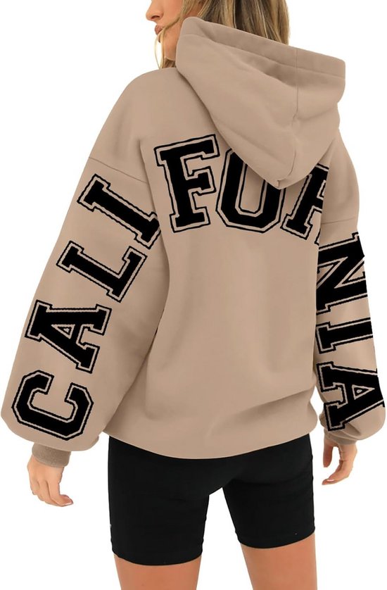 Vrouw in beige hoodie met zwarte letters op de mouwen en rug.