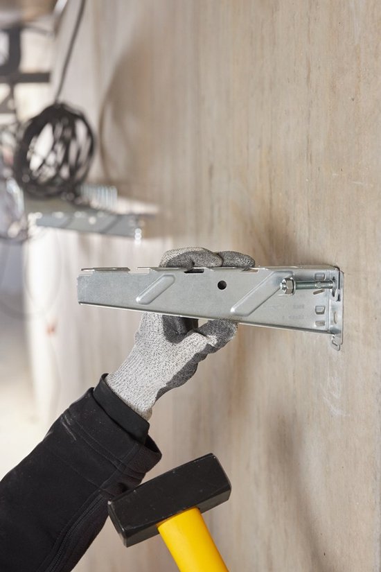 Een hand met een grijze werkhandschoen houdt een metalen montagebeugel vast, met een hamer naast de muur.