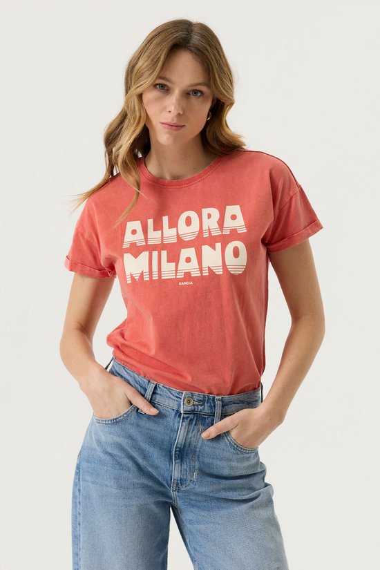 Femme portant un t-shirt corail "ALLORA MILANO GARCIA" et un jean, debout, mains dans les poches.