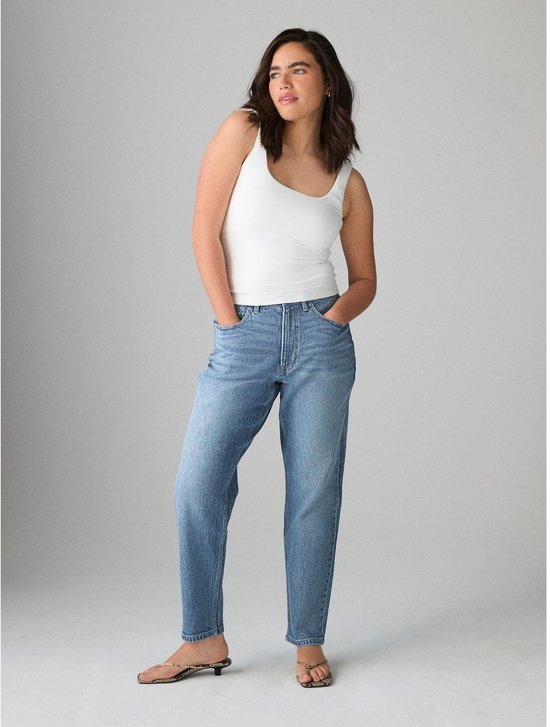 Model draagt een witte tanktop en blauwe, comfortabele mom jeans met hoge taille en lichte wassing. Staat met handen in de zakken.