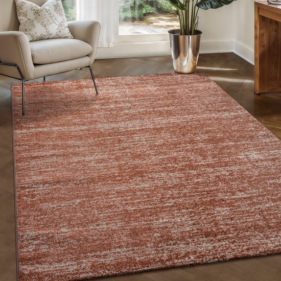 Terra-oranje laagpolig vloerkleed met een craquelé-achtig patroon in lichter grijs, in een woonkamer met een beige fauteuil.