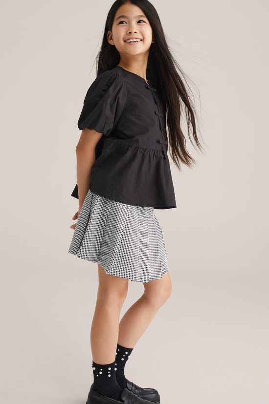 Meisje met lang zwart haar, in zwart pofmouwtje met strikjes en een geruite rok. Zwarte sokken met stippen en stoere schoenen.