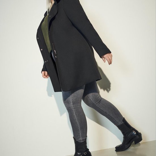 Vrouw in een zwarte lange jas met capuchon, olijfgroene top en grijze skinny jeans.