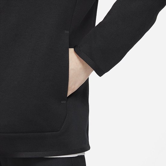 Detail van de hand van een jongen die in de zak van een zwart Nike Tech Fleece vest steekt.