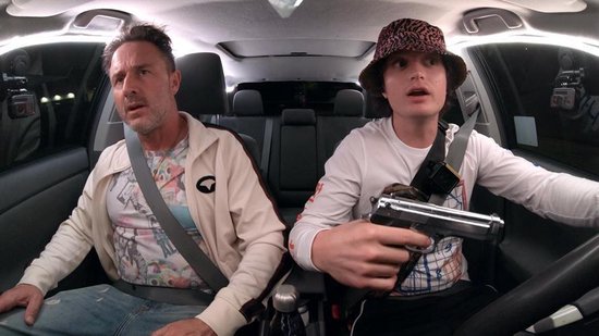 Twee mannen zitten in een auto. De passagier rechts draagt een bucket hat en houdt een pistool vast.