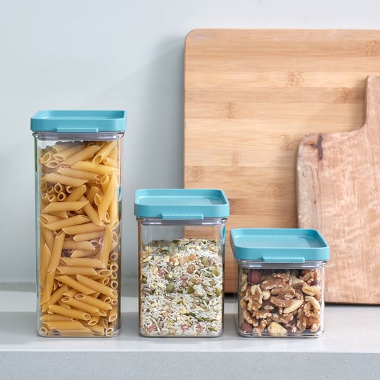 Trois boîtes de conservation transparentes avec des couvercles turquoise sur un plan de travail. Les boîtes sont remplies de pâtes penne, de riz aux graines et de noix.
