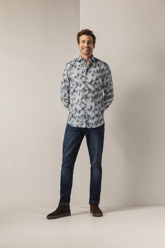Man met een glimlach draagt een lichtgrijs overhemd met botanische print en spijkerbroek.