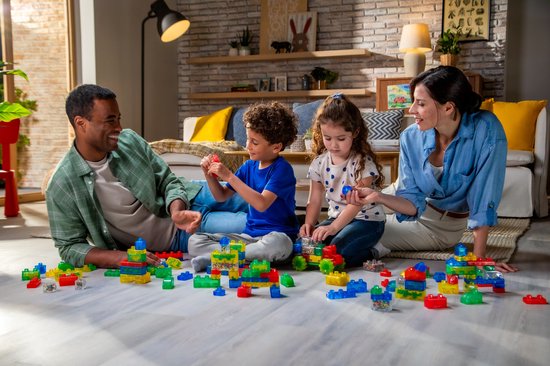 Een gezin (man, vrouw, jongen, meisje) zit op de grond en speelt met Jelly Blox bouwblokken.