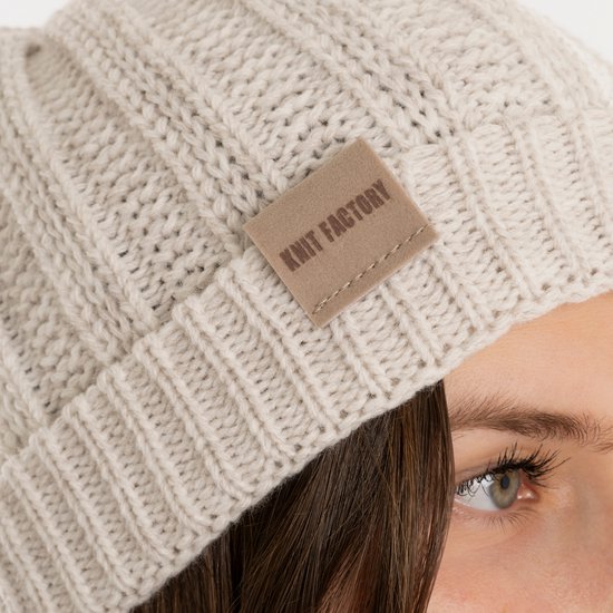 Close-up van een off-white gebreide muts met ribbelstructuur en beige label.