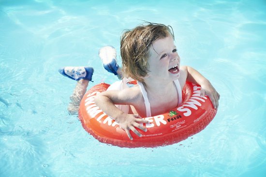 Lachend kind in rode SWIMTRAINER zwemband in blauw zwembadwater, met zwemvleugels aan de voeten.