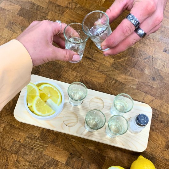 Deux mains lèvent des verres à shot pour trinquer, avec citron, sel et quatre verres à shot sur plateau en bois.