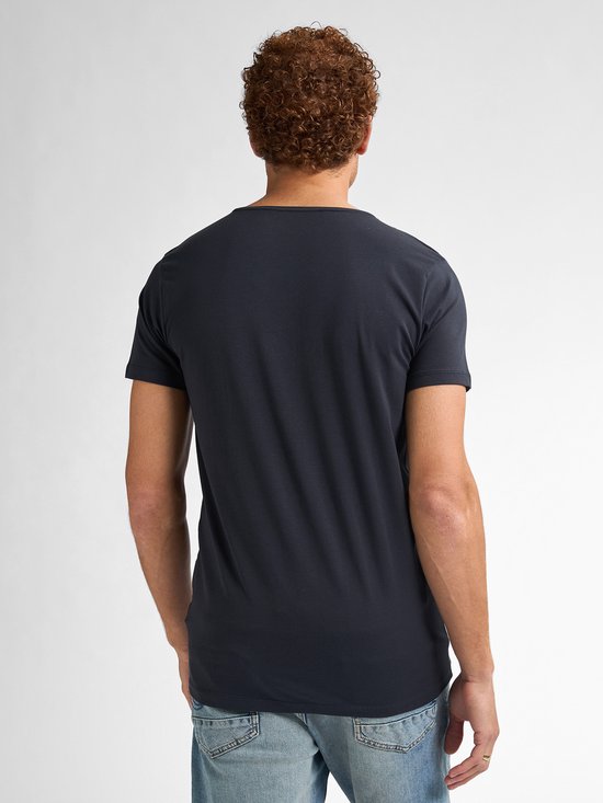 Achterkant van een man met krullend bruin haar die een donkerblauw V-hals T-shirt en een spijkerbroek draagt.