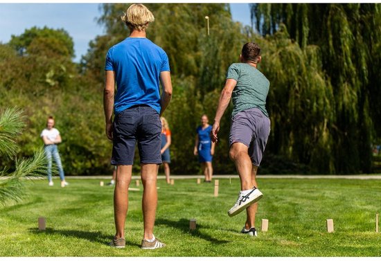Twee mannen spelen Kubb buiten op een grasveld, met andere spelers op de achtergrond.