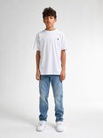 Jongen in een wit t-shirt en lichte, rechte spijkerbroek, staand op witte sneakers.
