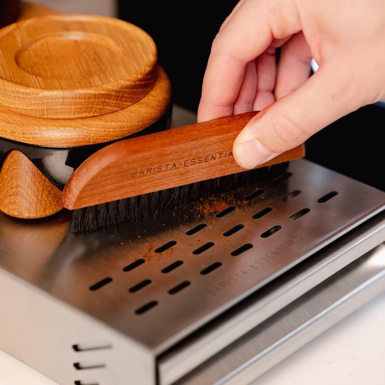 Een hand met een houten borstel met het opschrift 'BARISTA-ESSENTIALS' veegt koffiedik van een metalen rooster.