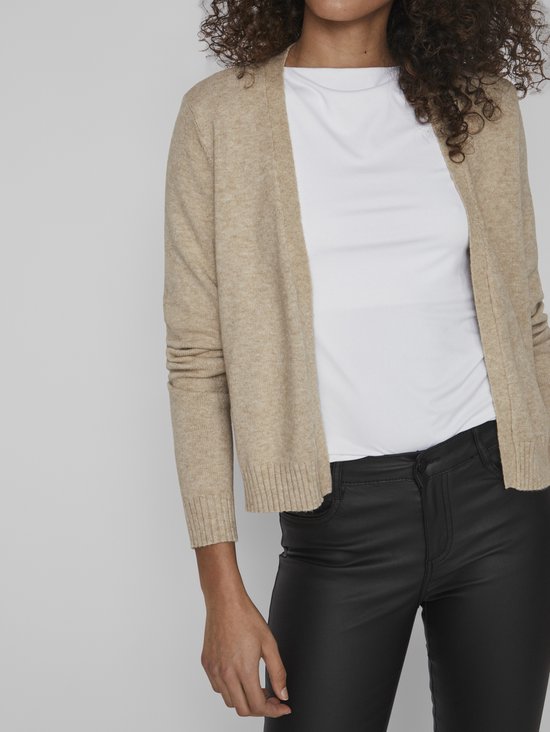 Lichtbeige gebreid vest met lange mouwen, gedragen over een wit shirt en zwarte leren broek.