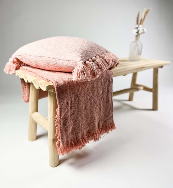 Zachtroze plaid met franjes en een geweven patroon, gestyled op een houten bankje met een bijpassend kussen.