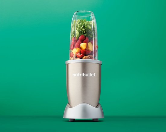 Nutribullet blender in champagnekleur op een groene achtergrond, met een beker gevuld met fruit.