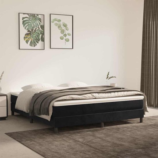 Een opgemaakt bed met een zwart boxspring frame, grijs plaid en twee groene planten aan de muur.