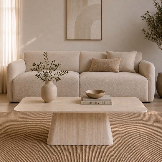 Table basse aspect travertin dans un salon avec un canapé beige, des coussins et un vase avec des branches.