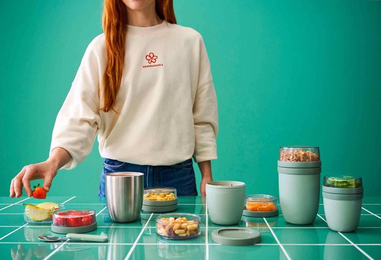 Een vrouw met een crèmekleurige sweater met rode bloem print staat achter verschillende Mepal lunchpots. De pot rechts bevat muesli, de pot midden met fruit, de pot links met groenten.