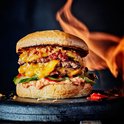 Een dubbele cheeseburger met gesmolten kaas, spek, augurk en rode pepers, met vlammen op de achtergrond.