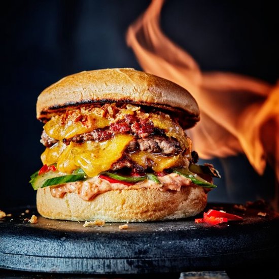 Een dubbele cheeseburger met gesmolten kaas, spek, augurk en rode pepers, met vlammen op de achtergrond.