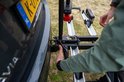 Handen bevestigen een zilverkleurige trekhaakfietsendrager aan de trekhaak van een auto, met een Nederlands kenteken zichtbaar.