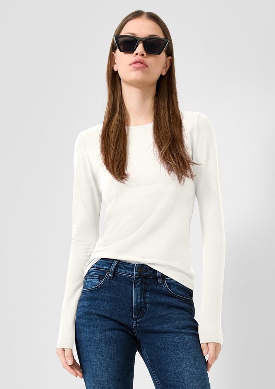 Model in een wit longsleeve shirt en donkerblauwe jeans, met haar los en een zwarte zonnebril op.