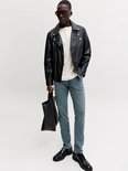 Man met zwarte bikerjas, crèmekleurige top, blauwe slim fit jeans, zonnebril en zwarte schoenen.