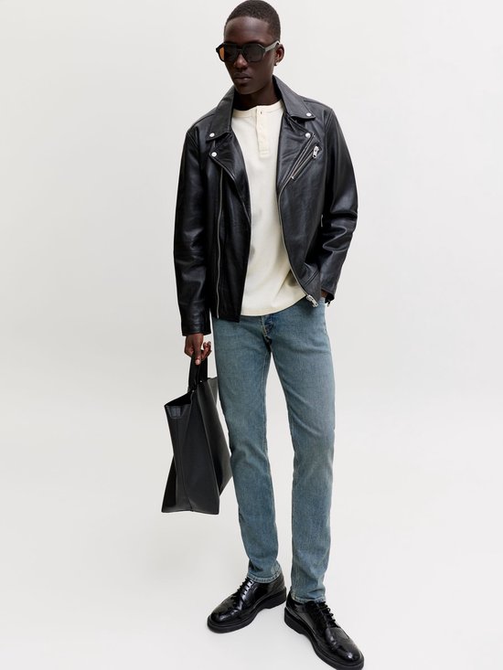 Man met zwarte bikerjas, crèmekleurige top, blauwe slim fit jeans, zonnebril en zwarte schoenen.